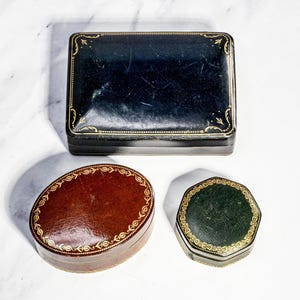 May include: A collection of three lidded boxes: a rectangular black box with gold filigree borders, an oval reddish-brown box with a gold leaf pattern around the edge, and a small octagonal dark green box featuring intricate gold decorative trim.