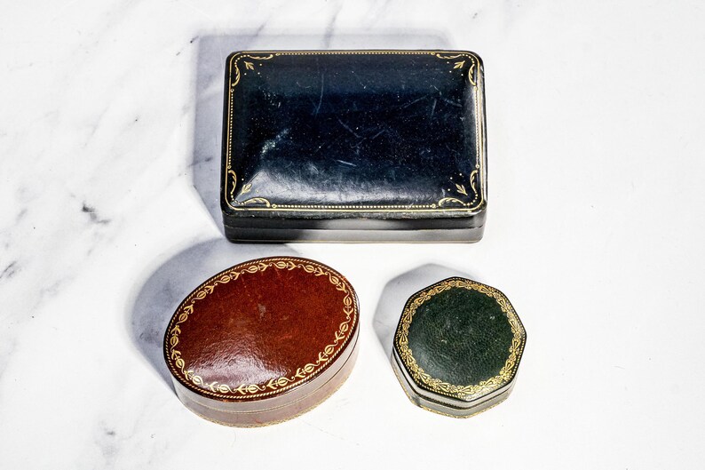 May include: A collection of three lidded boxes: a rectangular black box with gold filigree borders, an oval reddish-brown box with a gold leaf pattern around the edge, and a small octagonal dark green box featuring intricate gold decorative trim.