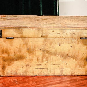 Puede incluir: Un cofre de madera rectangular con un acabado marr&oacute;n claro. El cofre tiene refuerzos de esquina y bisagras de metal. La veta de la madera es visible y el cofre parece envejecido. Se encuentra sobre una superficie marr&oacute;n rojiza.