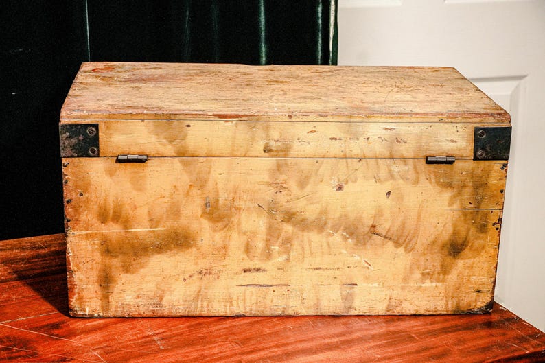 Puede incluir: Un cofre de madera rectangular con un acabado marr&oacute;n claro. El cofre tiene refuerzos de esquina y bisagras de metal. La veta de la madera es visible y el cofre parece envejecido. Se encuentra sobre una superficie marr&oacute;n rojiza.