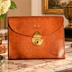 May include: A rectangular brown leather document case with a curved flap, featuring a central brass lock mechanism and the gold-embossed initials W. R. on the front. The leather has a textured, aged appearance with visible stitching along the edges.