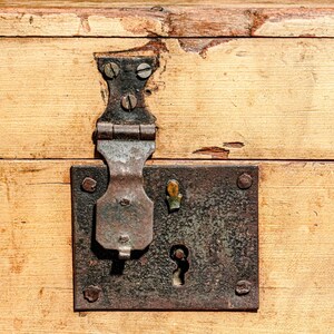 Puede incluir: Primer plano de una superficie de madera envejecida con un pestillo y cerradura de metal. El pestillo es de metal oscuro y envejecido, y la madera tiene un tono claro y c&aacute;lido con vetas visibles y algo de desgaste. La cerradura tiene un ojo de cerradura.