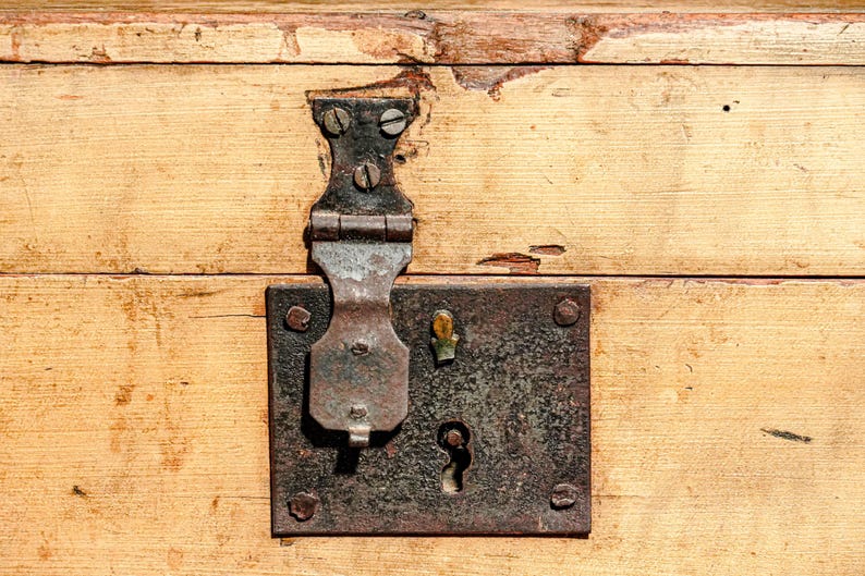 Puede incluir: Primer plano de una superficie de madera envejecida con un pestillo y cerradura de metal. El pestillo es de metal oscuro y envejecido, y la madera tiene un tono claro y c&aacute;lido con vetas visibles y algo de desgaste. La cerradura tiene un ojo de cerradura.