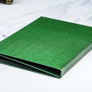 Puede incluir: Una caja rectangular verde con una superficie texturizada. La caja est&aacute; abierta, revelando un interior oscuro. La caja es probablemente para almacenamiento o presentaci&oacute;n. La caja est&aacute; sobre una superficie blanca.