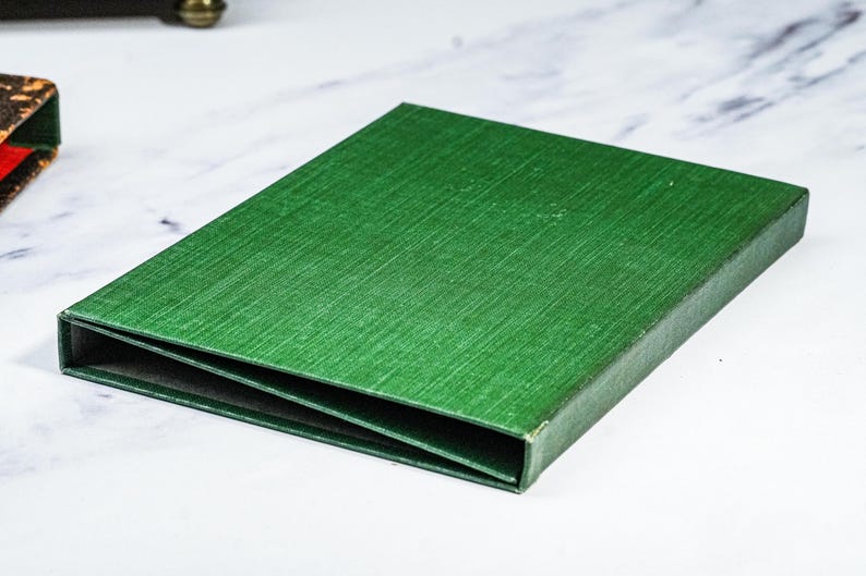 Puede incluir: Una caja rectangular verde con una superficie texturizada. La caja est&aacute; abierta, revelando un interior oscuro. La caja es probablemente para almacenamiento o presentaci&oacute;n. La caja est&aacute; sobre una superficie blanca.