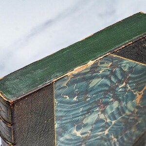 Puede incluir: Primer plano de un libro antiguo con lomo verde y cubierta veteada azul y verde. Los bordes del libro muestran signos de desgaste, con encuadernaci&oacute;n de cuero marr&oacute;n oscuro y esquina negra.