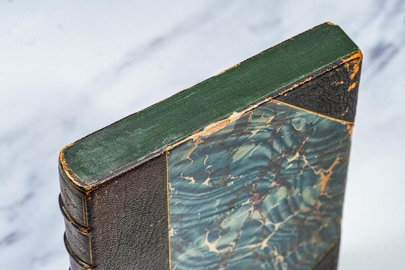 Puede incluir: Primer plano de un libro antiguo con lomo verde y cubierta veteada azul y verde. Los bordes del libro muestran signos de desgaste, con encuadernaci&oacute;n de cuero marr&oacute;n oscuro y esquina negra.