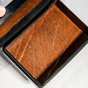 May include: A rectangular box with a black exterior and a textured, light brown interior lining. The lid is partially open, revealing the contrasting dark outer frame and the tan, grained surface of the base interior. The box has a hinged design.