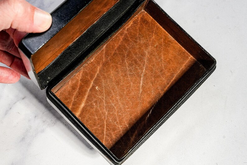 May include: A rectangular box with a black exterior and a textured, light brown interior lining. The lid is partially open, revealing the contrasting dark outer frame and the tan, grained surface of the base interior. The box has a hinged design.