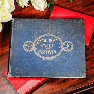 May include: A vintage book titled "American Art and Artists" with gold lettering on a blue cover. The book is resting on a dark wooden surface, with a red book partially visible beneath it. White roses are in the background.