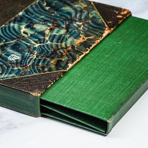 Puede incluir: Un libro antiguo con una cubierta jaspeada en verde azulado y marr&oacute;n, y una caja verde. La cubierta del libro tiene un aspecto texturizado y envejecido. La caja es de color verde oscuro liso, proporcionando un estuche protector para el libro.