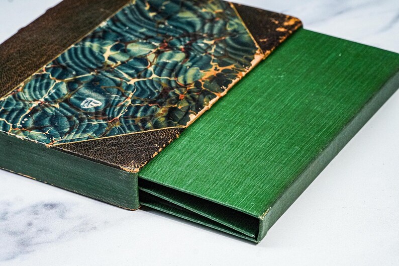Puede incluir: Un libro antiguo con una cubierta jaspeada en verde azulado y marr&oacute;n, y una caja verde. La cubierta del libro tiene un aspecto texturizado y envejecido. La caja es de color verde oscuro liso, proporcionando un estuche protector para el libro.