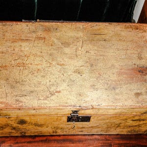 Puede incluir: Una caja de madera rectangular con un aspecto envejecido y desgastado. La caja es de color marr&oacute;n claro con ara&ntilde;azos y desgaste visibles, y presenta refuerzos de esquina y un pestillo de metal negro. La caja est&aacute; sobre una superficie de madera oscura.