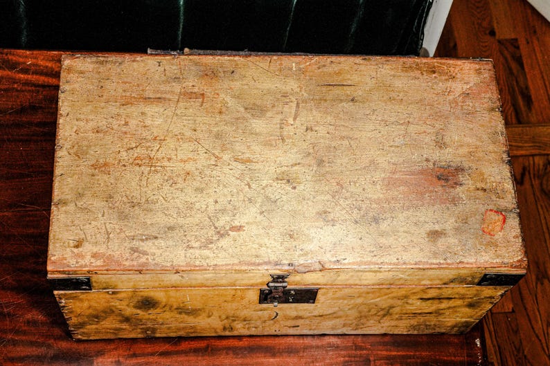 Puede incluir: Una caja de madera rectangular con un aspecto envejecido y desgastado. La caja es de color marr&oacute;n claro con ara&ntilde;azos y desgaste visibles, y presenta refuerzos de esquina y un pestillo de metal negro. La caja est&aacute; sobre una superficie de madera oscura.