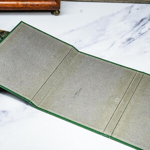 Puede incluir: Un libro abierto, con bordes verdes e interior gris, se encuentra junto a un libro vintage con una cubierta estampada. El libro abierto est&aacute; doblado en tres secciones. El fondo es una superficie blanca con vetas grises.