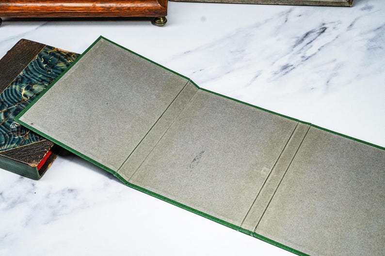 Puede incluir: Un libro abierto, con bordes verdes e interior gris, se encuentra junto a un libro vintage con una cubierta estampada. El libro abierto est&aacute; doblado en tres secciones. El fondo es una superficie blanca con vetas grises.