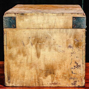 Puede incluir: Una caja de madera envejecida con refuerzos de esquina met&aacute;licos. La caja tiene forma rectangular con tapa y muestra signos de desgaste, mostrando una est&eacute;tica vintage. La madera tiene un tono dorado c&aacute;lido con manchas y ara&ntilde;azos m&aacute;s oscuros.