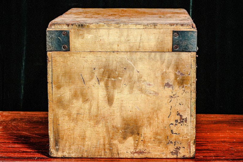 Puede incluir: Una caja de madera envejecida con refuerzos de esquina met&aacute;licos. La caja tiene forma rectangular con tapa y muestra signos de desgaste, mostrando una est&eacute;tica vintage. La madera tiene un tono dorado c&aacute;lido con manchas y ara&ntilde;azos m&aacute;s oscuros.