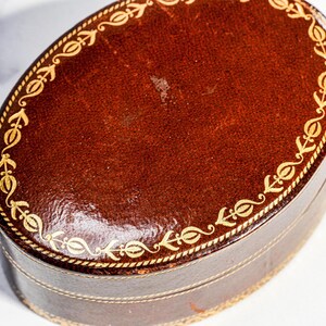 May include: An oval-shaped dark brown leather box featuring a gold-coloured decorative border with a repeating floral pattern along the edge of the lid and the base. The surface of the leather has a smooth, polished finish with visible texture.