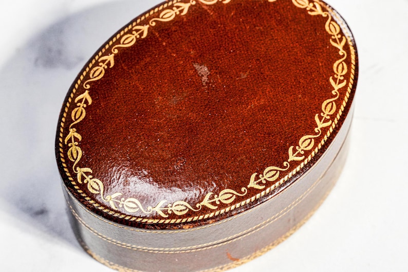 May include: An oval-shaped dark brown leather box featuring a gold-coloured decorative border with a repeating floral pattern along the edge of the lid and the base. The surface of the leather has a smooth, polished finish with visible texture.