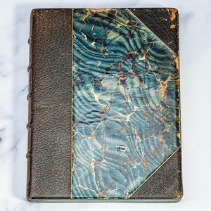 Puede incluir: Libro antiguo con lomo y esquinas de cuero marr&oacute;n oscuro. La cubierta presenta un dise&ntilde;o veteado en tonos turquesa, azul y crema. Los bordes del libro muestran signos de desgaste, lo que indica su antig&uuml;edad. Una pieza cl&aacute;sica para coleccionistas.