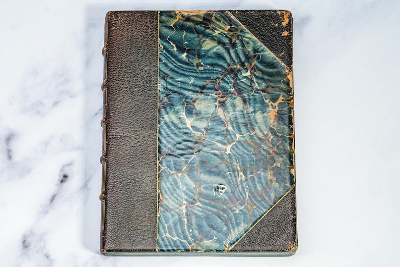 Puede incluir: Libro antiguo con lomo y esquinas de cuero marr&oacute;n oscuro. La cubierta presenta un dise&ntilde;o veteado en tonos turquesa, azul y crema. Los bordes del libro muestran signos de desgaste, lo que indica su antig&uuml;edad. Una pieza cl&aacute;sica para coleccionistas.