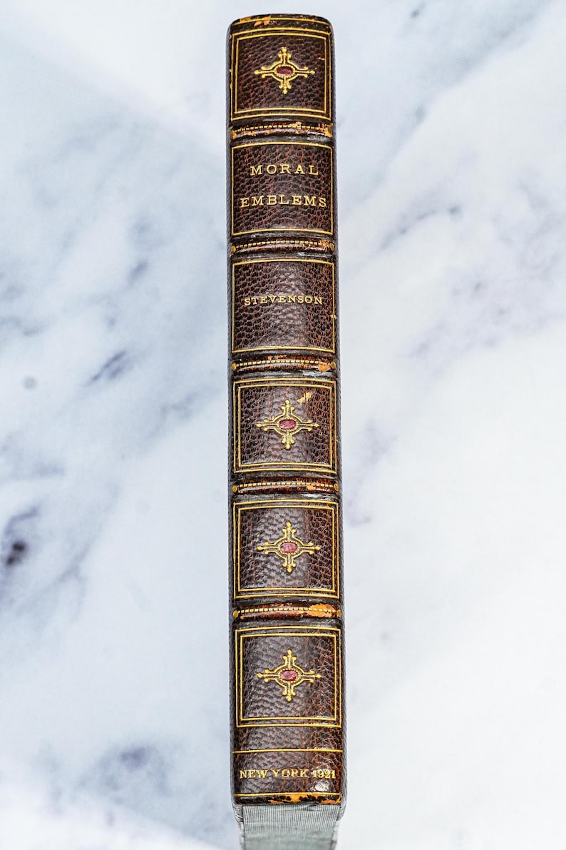Puede incluir: Lomo de libro antiguo con el t&iacute;tulo "MORAL EMBLEMS" y el autor "STEVENSON" en letras doradas. La cubierta de cuero marr&oacute;n presenta detalles dorados y un emblema rojo. El libro es de Nueva York, 1921.