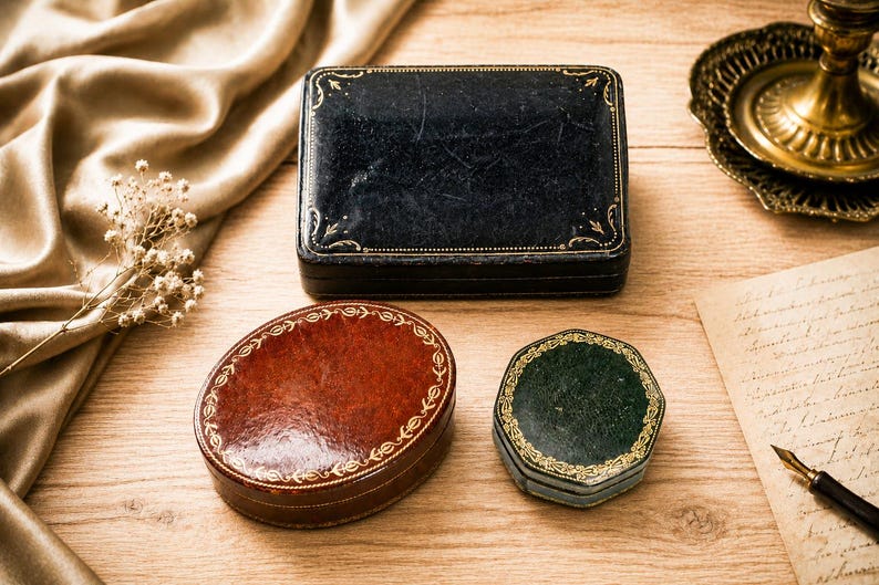 May include: Three small storage boxes: a rectangular black box, an oval reddish-brown box, and a hexagonal dark green box. Each box features a smooth, textured surface with intricate gold-coloured decorative filigree patterns along the edges.