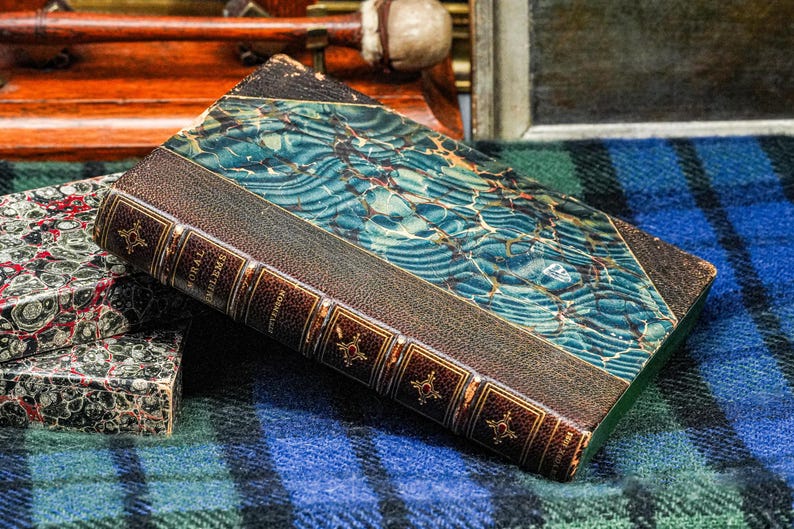 Puede incluir: Libro antiguo con cubierta azul y verde veteada y lomo de cuero marr&oacute;n. El lomo tiene letras doradas y elementos decorativos. El libro descansa sobre una superficie a cuadros azul y verde, con otro libro visible.