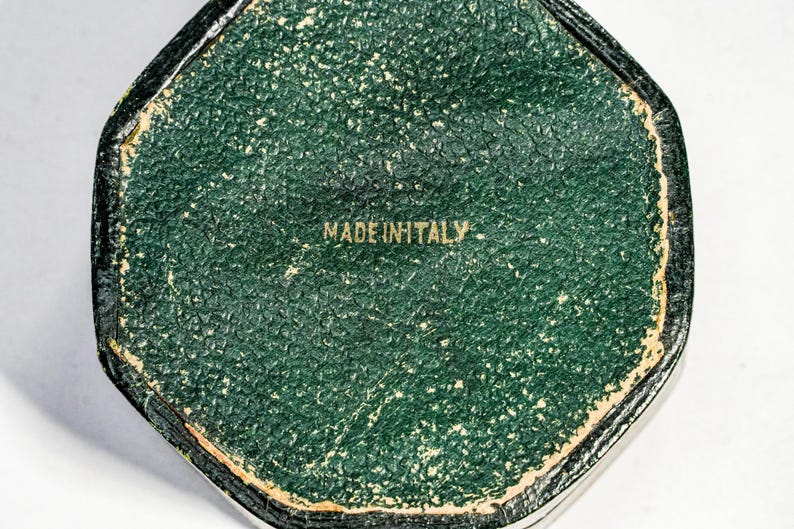 May include: A hexagonal object with a textured dark green surface featuring a gold-coloured border and the text MADE IN ITALY printed in the centre. The object has a worn appearance with visible signs of age along the edges and the green surface.