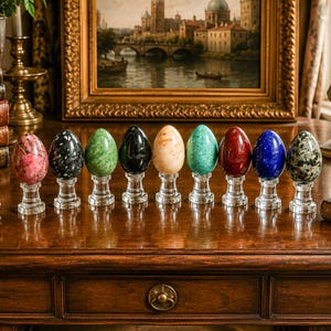 May include: A collection of nine polished stone eggs in various colors including pink, black, green, cream, red, and blue, each mounted on a clear glass pedestal base. The eggs display unique natural mineral patterns and textures throughout.