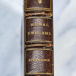 Puede incluir: Primer plano del lomo de un libro antiguo. El libro se titula "MORAL EMBLEMS" de Stevenson, con letras doradas sobre una cubierta de cuero marr&oacute;n oscuro. La cubierta tiene adornos y elementos decorativos dorados.