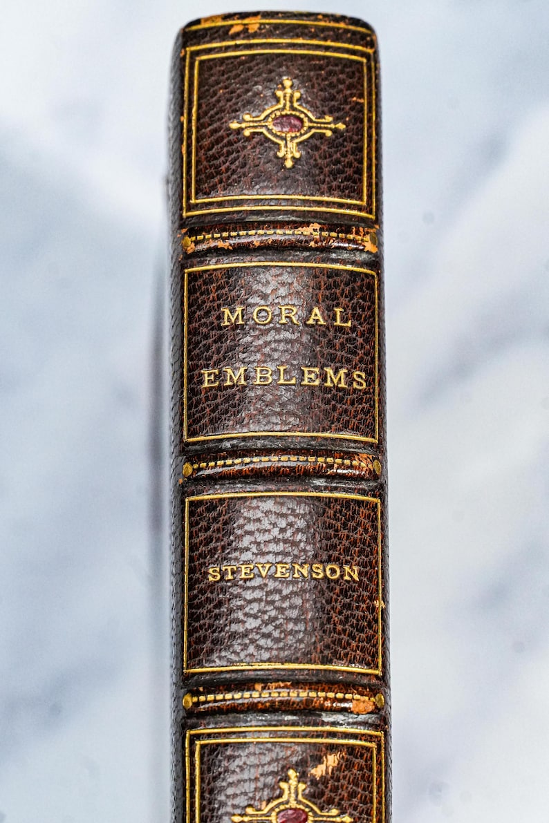 Puede incluir: Primer plano del lomo de un libro antiguo. El libro se titula "MORAL EMBLEMS" de Stevenson, con letras doradas sobre una cubierta de cuero marr&oacute;n oscuro. La cubierta tiene adornos y elementos decorativos dorados.