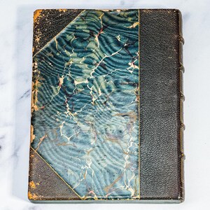 Puede incluir: Un libro antiguo con una cubierta marmolada azul y blanca y un lomo de cuero negro. La cubierta presenta un patr&oacute;n &uacute;nico con tonos de azul y blanco, y el lomo es oscuro. Los bordes del libro muestran signos de desgaste, lo que sugiere su antig&uuml;edad.