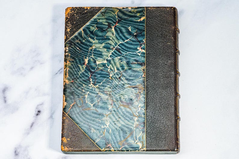 Puede incluir: Un libro antiguo con una cubierta marmolada azul y blanca y un lomo de cuero negro. La cubierta presenta un patr&oacute;n &uacute;nico con tonos de azul y blanco, y el lomo es oscuro. Los bordes del libro muestran signos de desgaste, lo que sugiere su antig&uuml;edad.