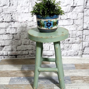 Pode incluir: Um banco redondo de madeira verde-claro com acabamento desgastado e quatro pernas ligadas por suportes horizontais. No topo, um vaso de cerâmica com padrão floral azul, verde e branco contém uma planta verde com caules finos.