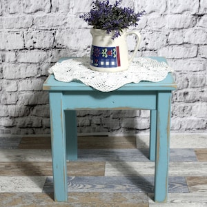 May include: A small, square, turquoise-painted wooden side table with distressed edges. A white crocheted doily and a cream-colored pitcher with a floral design and purple faux flowers sit on top. The table is on a wood-look floor.