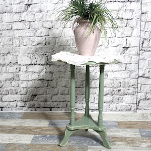 Könnte beinhalten: Kleiner, salbeigrüner Pflanzenständer mit runder Oberseite und drei Beinen. Eine rosa Vase mit Grünzeug steht auf einer weißen Spitzendecke. Der Ständer ist mit einem Used-Look lackiert, und der Hintergrund ist eine weiße Backsteinmauer.