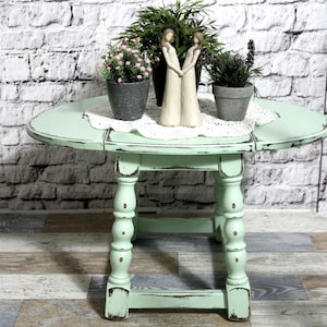 Può includere: Un tavolo rotondo a ribalta color verde chiaro con finitura usurata e gambe tornite. Sul tavolo si trovano un centrino di pizzo bianco, due piccole piante in vaso grigio e una statuetta alta color crema di due figure che si tengono.