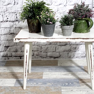 Op de afbeelding: Een kleine rechthoekige tafel met een verweerde witte afwerking en vier taps toelopende poten. Op het tafelblad staan vier potplanten: een grote groene plant in een donkere metalen pot, twee kleine grijze potten met bloemen en een groene kan.