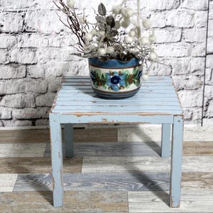 May include: A small, square, pale blue wooden table with a distressed finish. A ceramic pot with a floral design and decorative branches sits on top. The table has four legs and a slatted top.