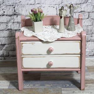 Op de afbeelding: Een roze nachtkastje met twee lades en een decoratief bovenblad. De lades zijn wit met roze knoppen. Het nachtkastje is versierd met een wit kanten kleedje, een vaasje met paarse bloemen en twee engelenbeeldjes.