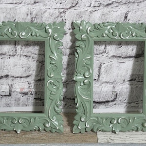 Könnte beinhalten: Zwei verzierte, salbeigrüne Bilderrahmen mit detaillierten Schnörkeln und weißen Akzenten. Die rechteckigen Rahmen haben ein klassisches, Vintage-Design, ideal für Fotos oder Kunstwerke. Sie stehen vor einer strukturierten, hellfarbigen Backsteinmauer.