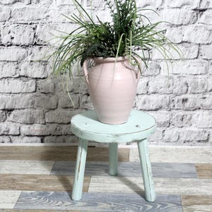 May include: A light pink ceramic vase with two handles, filled with greenery, sits on a small, round, three-legged, distressed, light blue wooden stool. The background is a white brick wall.