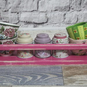 Könnte beinhalten: Ein rosa, verwittertes Holzregal zeigt eine Sammlung von Teetassen und kleinen Behältern. Die Teetassen sind mit floralen Mustern verziert, und die Behälter haben Deckel. Das Regal hat ein abgenutztes, Vintage-Aussehen.