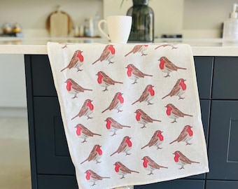 May include: A white kitchen towel featuring a repeating pattern of brown and red robins. The birds are depicted in a detailed illustration, with their red breasts and brown wings. The towel is folded over a dark blue cabinet.