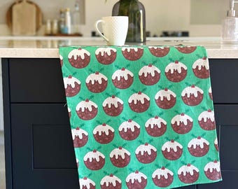 May include: A green kitchen towel with a repeating pattern of Christmas pudding illustrations. Each pudding is brown with white icing, a red berry, and holly leaves. The towel is draped over a dark blue cabinet.