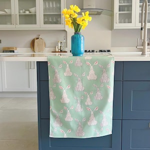 May include: A green kitchen towel with a white rabbit pattern. The towel is hanging on a kitchen counter with a vase of yellow flowers in the background.