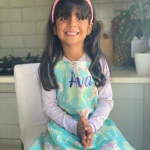 May include: A young girl wearing a light blue apron with a green and white animal print and the name "Ava" embroidered in purple. The apron has a pocket and adjustable straps.