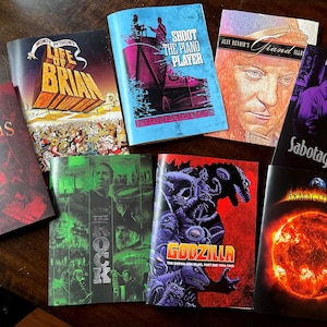 May include: A collection of vintage movie posters, including "Spartacus," "Monty Python's Life of Brian," and "Godzilla." The posters feature colorful illustrations and text, showcasing classic film titles and artwork. The posters are arranged on a dark wooden surface.